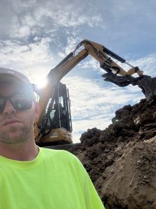 Nicholas wearing a neon yellow shirt and sunglasses stands in front of construction equipment at a dirt worksite.