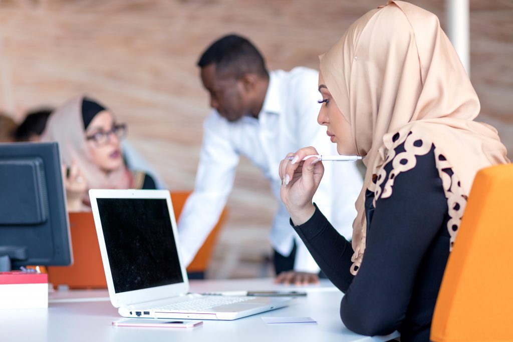 A woman wearing a hijab works on a laptop while a job coach and another participant talk in the background.
