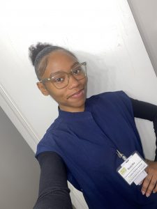 Denysha smiling in her CNA uniform and glasses while standing indoors.
