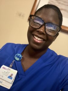 Ebony smiles while wearing blue nursing scrubs and glasses.
